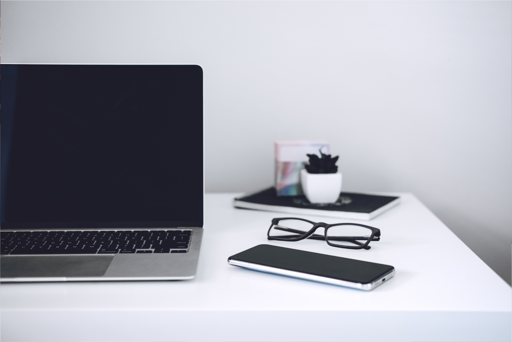 An organized workspace featuring a laptop with the screen off, a smartphone, an open book, a pen, and reading glasses. The setup invites readers to take notes while exploring blog content